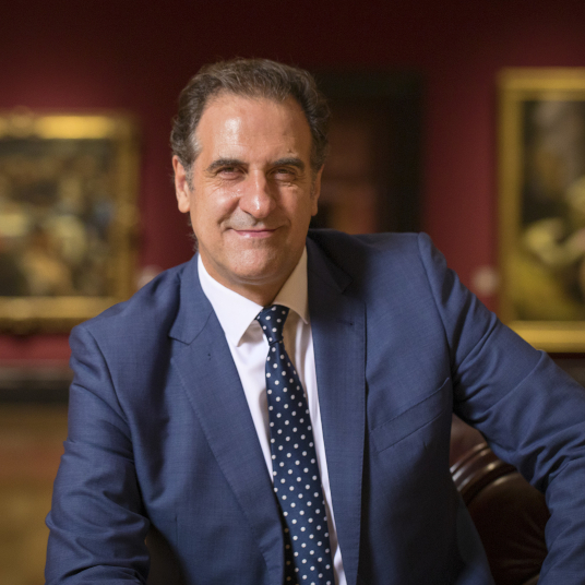 Photograph of Gabriele Finaldi: a man with dark hair in a blue suit and tie sitting in an art gallery.