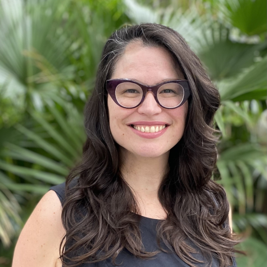 Photographic headshot of Leslie Geddes: a smiling woman with dark hair and purple glasses with leafy branches behind her. 